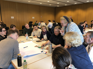 Eine vielfältige Gruppe von Menschen arbeitet an Tischen in einem Workshop zusammen, wobei eine Frau aktiv eine Idee präsentiert.