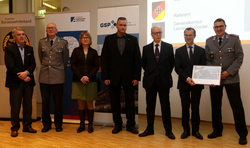 Gruppenfoto von sieben Personen, darunter Militärpersonal, vor Veranstaltungsbannern, einer hält ein gerahmtes Bild.