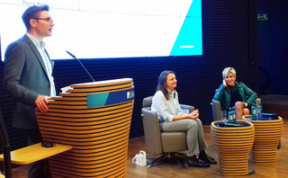 Paneldiskussion mit einem Mann, der an einem Rednerpult spricht, und zwei Frauen, die in Sesseln sitzen und zuhören.