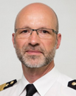 Porträt eines glatzköpfigen Mannes mit Brille und Bart, der ein weißes Uniformhemd mit schwarzer Krawatte und Schulterklappen trägt.