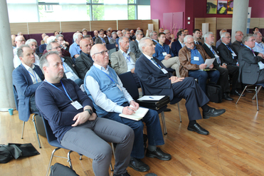 Eine Gruppe von Männern sitzt in Reihen in einem hellen Konferenzraum und schaut aufmerksam nach vorne.
