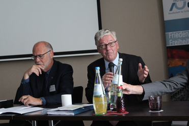 Drei Männer bei einer Konferenz: Einer mit grauen Haaren spricht ins Mikrofon, ein bärtiger Mann hört zu, der Arm einer dritten Person ist sichtbar.