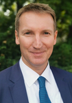 Porträt eines lächelnden Mannes im blauen Anzug und Krawatte vor verschwommenem grünem Hintergrund.