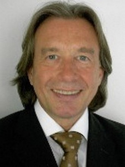 Ein lächelnder Mann mit mittellangem braunem Haar trägt einen dunklen Anzug, ein weißes Hemd und eine gemusterte Krawatte.