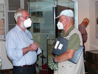Zwei Männer mit Masken sprechen in einem Museum, hinter ihnen eine Tafel über den STALAG Friedhof I Alliiertenfriedhof.