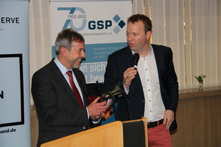 Ein Mann im Anzug erhält ein Geschenk von einem Mann mit Mikrofon an einem Podium bei einer GSP-Veranstaltung.