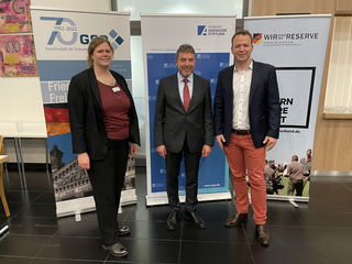 Drei Personen, eine Frau und zwei Männer, lächeln vor Bannern der GSD, Konrad-Adenauer-Stiftung und dem Reservistenverband.