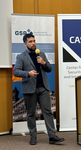 Ein bärtiger Mann im Sakko spricht in ein gelbes Mikrofon, stehend zwischen zwei Event-Bannern, eines für 'GSP' und eines für 'CAS'.