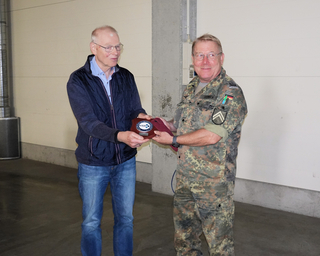 Zwei Männer, einer in Militäruniform, der andere in Zivil, tauschen lächelnd eine GSP-Plakette aus.