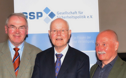 Drei lächelnde ältere Männer, zwei in Anzügen und einer im Blazer, stehen vor einem Banner der 'Gesellschaft für Sicherheitspolitik'.