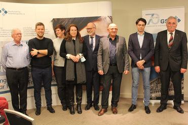 Neun Personen, Männer und Frauen, stehen in einer Reihe vor Bannern der Gesellschaft für Sicherheitspolitik (GSP) bei einer Veranstaltung.