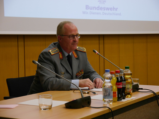 Mann in Bundeswehr-Uniform bei Präsentation am Tisch mit Mikrofon und Getränken. Bundeswehr-Logo im Hintergrund.