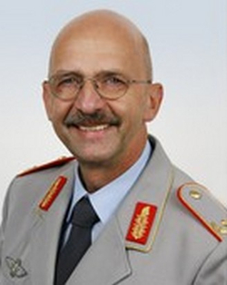 Lächelnder Glatzkopf mit Schnurrbart und Brille in grauer Militäruniform mit roten und goldenen Abzeichen.