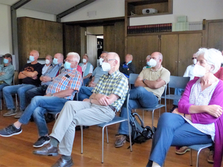 Eine vielfältige Gruppe von Erwachsenen mit Gesichtsmasken sitzt aufmerksam bei einer Indoor-Veranstaltung oder Vortrag.