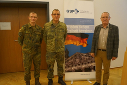 Drei Männer, zwei in Bundeswehruniform, einer in Zivil, posieren vor einem Banner der Gesellschaft für Sicherheitspolitik (GSP).