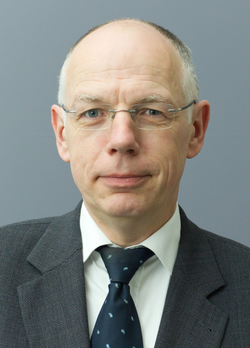 Professionelles Porträt eines Mannes mit grauen Haaren, Brille, dunklem Anzug und gemusterter blauer Krawatte.
