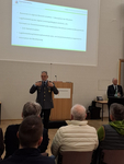 Ein uniformierter Offizier präsentiert auf einer Konferenz, dahinter eine Folie zu Militärkrankenhausbetrieb und Katastrophenschutz.