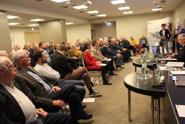 Zahlreiche Personen sitzen in einem Konferenzraum und lauschen zwei Sprechern bei einer Präsentation oder einem Meeting.