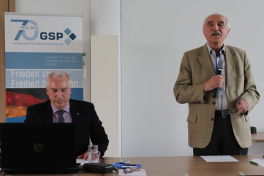 Ein älterer Mann spricht ins Mikrofon, ein anderer sitzt am Laptop. GSP-Banner und deutsche Flagge sind im Hintergrund.