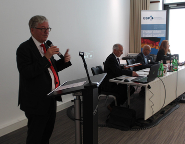 Ein Mann im Anzug spricht an einem Rednerpult mit Mikrofon auf einer Konferenz mit anderen Teilnehmern und GSP-Banner.