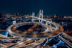 Nachtaufnahme einer Stadt mit spiralförmiger Autobahnkreuzung, Lichtspuren von Fahrzeugen und einer beleuchteten Brücke.