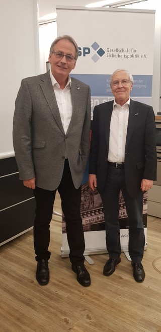 Zwei Männer in Sakkos und Brillen lächeln vor einem Banner der Gesellschaft für Sicherheitspolitik (GSP).
