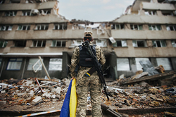 Soldat mit ukrainischer Flagge vor einem schwer beschädigten Gebäude, umgeben von Trümmern, Symbol für Krieg und Zerstörung.