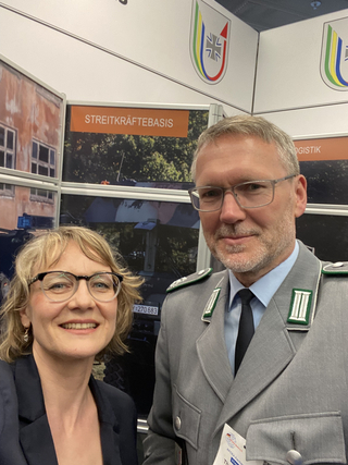 Zwei lächelnde Personen, eine Frau und ein Bundeswehrsoldat, vor einem Stand der Streitkräftebasis.