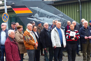 Eine Gruppe älterer Erwachsener steht vor einem Flugzeugheck mit deutscher Flagge und Biplan-Gemälden.
