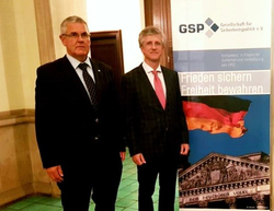 Zwei Männer in Anzügen stehen vor einem GSP-Banner mit deutscher Flagge und Reichstagsgebäude, das Sicherheit und Freiheit fördert.