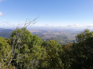 Panoramablick über ein Tal mit Dorf und Feldern, umrahmt von grünen Bäumen, unter blauem Himmel.