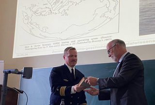 Ein Marineoffizier in Uniform erhält ein Buch von einem Mann im Anzug bei einer Präsentation mit einer Karte im Hintergrund.