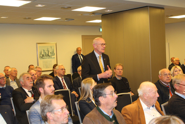 Ein Mann im Anzug spricht vor einem Publikum, hauptsächlich ältere Männer, in einem Konferenzraum.