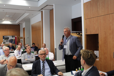 Ein Mann mit Mikrofon spricht vor einer Gruppe von Teilnehmern in einem Konferenzraum. Andere sitzen an Tischen und hören zu.