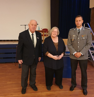 Ein älterer Mann, eine ältere Frau und ein Soldat in deutscher Uniform stehen gemeinsam auf einer Bühne.
