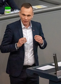 Ein Mann mit kurzen Haaren im dunklen Anzug und weißen Hemd spricht an einem Rednerpult und gestikuliert.