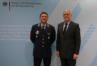 Ein deutscher Offizier in Uniform und ein Mann im Anzug lächeln vor der Wand des Bundespresseamtes.