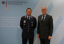Ein deutscher Offizier in Uniform und ein Mann im Anzug lächeln vor der Wand des Bundespresseamtes.