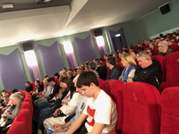 Ein vielfältiges Publikum jeden Alters sitzt auf roten Stühlen in einem Theater mit lila und grünen Wänden.