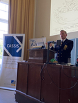 Ein Marineoffizier in Uniform präsentiert von einem Pult, mit CASSIS-Banner und einer Karte auf einer Leinwand.