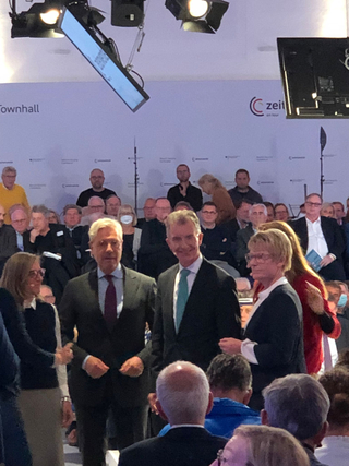 Drei Männer und eine Frau stehen auf der Bühne einer "Townhall"-Veranstaltung mit Publikum. Ein "Zeit en tour"-Logo ist sichtbar.