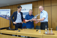 Zwei Männer überreichen sich ein verpacktes Geschenk bei einer Veranstaltung, ein dritter Mann steht daneben. Banner von 'Bonifatiushaus' und 'GSP' sind sichtbar.