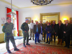 Zwei uniformierte Soldaten und eine Gruppe Zivilisten stehen auf einem roten Teppich in einem formellen Raum mit roten Vorhängen.