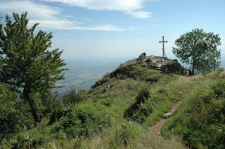 Gipfelkreuz auf einem Berg mit Wanderweg, weitem Talblick und grüner Vegetation unter blauem Himmel.