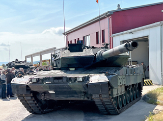 Ein Leopard 2 Kampfpanzer auf Ausstellung. Menschen betrachten ihn vor einem roten Gebäude. Ein weiterer Panzer im Hintergrund.