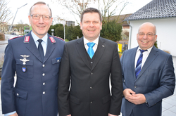 Drei lächelnde Männer, einer in dunkler Uniform und zwei in Anzügen, stehen draußen.