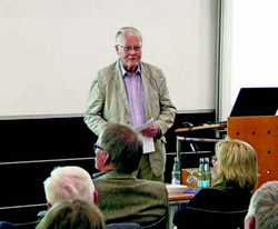 Ein älterer Mann mit Brille und Jackett lächelt, während er eine Präsentation vor einem Publikum hält, Dokumente in der Hand.