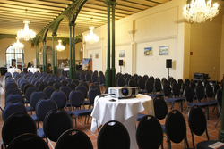 Konferenzraum mit blauen Stuhlreihen, Projektor, Kronleuchtern, grünen Metallbögen und hoher Decke.