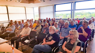 Eine große Gruppe älterer Menschen sitzt in einem hellen Raum mit Fenstern und nimmt an einer Veranstaltung teil.
