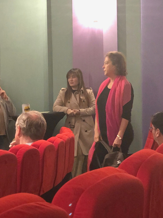 Zwei Frauen in einem Auditorium mit roten Sitzen. Eine trägt einen rosa Schal, die andere einen Trenchcoat, nahe einem 'Help'-Becher.
